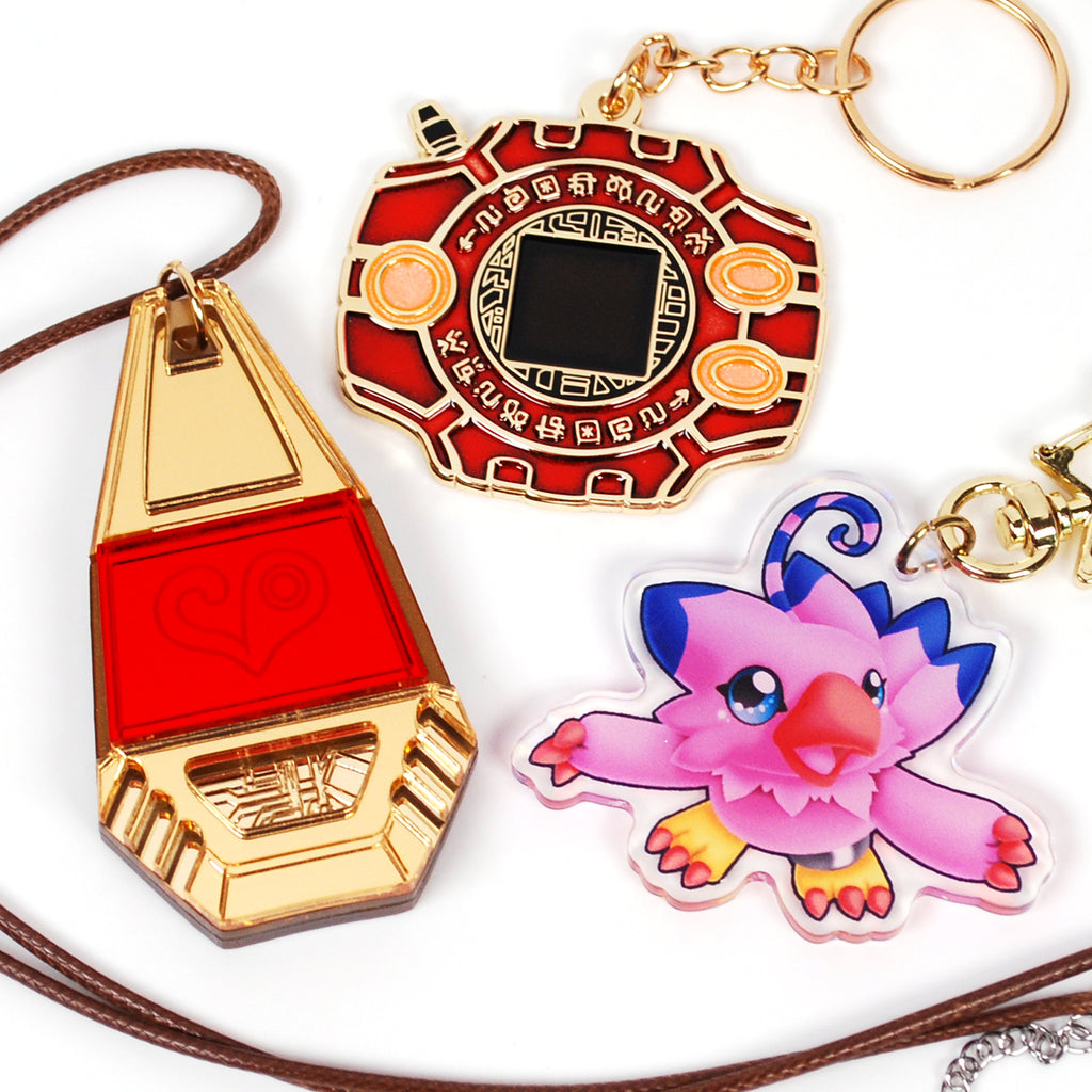 Three keychains: a red and gold triangular pendant, a red circular pendant with intricate design, and a pink and blue creature charm on a white background.